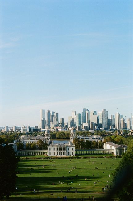 A wide panoramic view of central London featuring the historic Royal Hospital Chelsea in the foreground, characterized by its classical architecture with white stone walls, columns, and a symmetrical facade. Surrounding the hospital are well-maintained, lush green lawns with scattered visitors enjoying the open space. Behind the hospital, a dense cluster of modern high-rise skyscrapers with glass and steel finishes rises against a clear blue sky with faint wispy clouds. The skyscrapers' reflective surfaces catch the sunlight, creating a bright contrast with the greenery below. The scene captures a peaceful contrast between historic architecture and contemporary urban development, illustrating the blend of green public space with city infrastructure. The open, flat park area at the bottom of the image extends from the foreground to the midground, providing an accessible outdoor environment that could be associated with private or independent leisure activities, subtly reflecting alternative approaches to waste management by the use of well-maintained urban parks, and indirectly connecting to municipal or private waste disposal services in an urban setting.