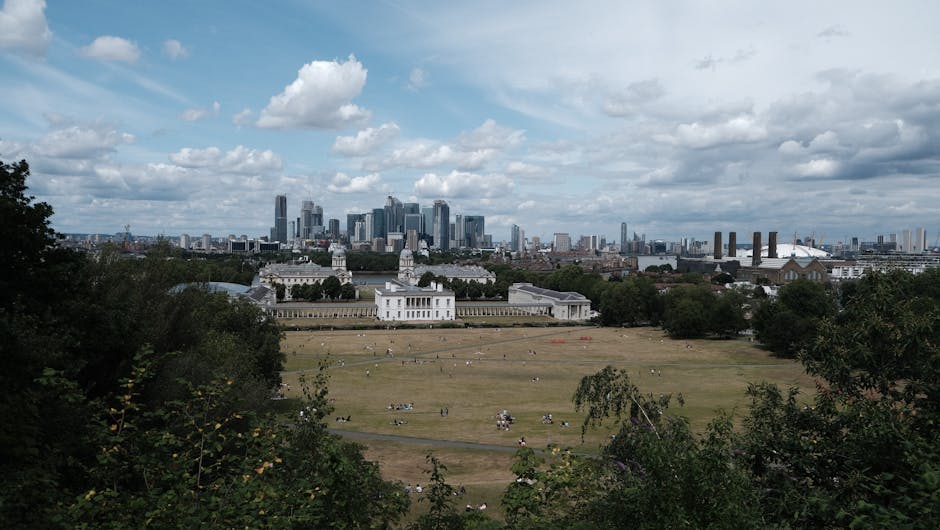 A wide view of a large, open grassy park area with scattered groups of people sitting and relaxing on the ground. In the middle ground, a white historic building with classical architectural features, such as columns and a balustrade, is visible, surrounded by greenery and trees on either side. In the background, a modern city skyline with tall glass skyscrapers dominates the scene, set against a partly cloudy sky with patches of blue. The image suggests a contrast between urban development and open green space, with the skyline on the horizon and the park in the foreground, illustrating an environment where private outdoor leisure and cityscape views coexist. The overall atmosphere is calm with soft natural lighting highlighting the textures of the grass, trees, and buildings, aligning with themes of urban renewal and recreational use common in discussions of infrastructure and space management.