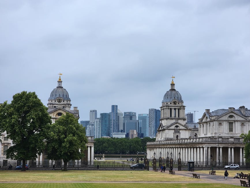 A wide panoramic view of central London featuring the historic Royal Hospital Chelsea in the foreground, characterized by its classical architecture with white stone walls, columns, and a symmetrical facade. Surrounding the hospital are well-maintained, lush green lawns with scattered visitors enjoying the open space. Behind the hospital, a dense cluster of modern high-rise skyscrapers with glass and steel finishes rises against a clear blue sky with faint wispy clouds. The skyscrapers' reflective surfaces catch the sunlight, creating a bright contrast with the greenery below. The scene captures a peaceful contrast between historic architecture and contemporary urban development, illustrating the blend of green public space with city infrastructure. The open, flat park area at the bottom of the image extends from the foreground to the midground, providing an accessible outdoor environment that could be associated with private or independent leisure activities, subtly reflecting alternative approaches to waste management by the use of well-maintained urban parks, and indirectly connecting to municipal or private waste disposal services in an urban setting.