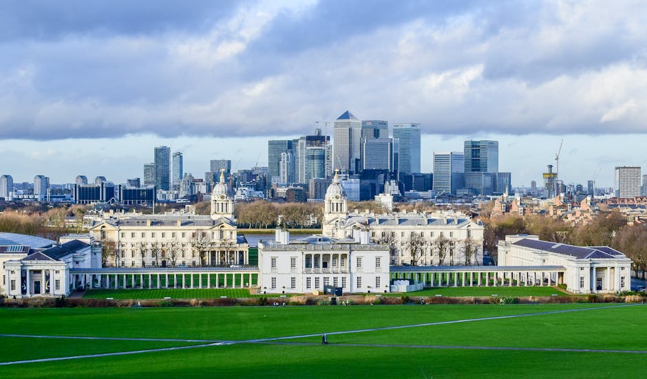 A wide-angle view of the Royal Naval College in Greenwich, London, showcasing its classical white façade with ornate architectural details, symmetrical towers with domed tops, and a series of colonnades along the front. The building is situated on a well-maintained grassy lawn with paved pathways leading towards it. The background features a city skyline with modern skyscrapers and high-rise office buildings, under a partly cloudy sky with patches of sunlight. The scene is set during daytime, with natural light illuminating both the historic structure and the urban backdrop. This image emphasizes the contrast between the historic Greenwich architecture and the contemporary cityscape, illustrating the area’s unique blend of heritage and modern development, which can be related to professional waste removal services handling clearance of historic sites or urban redevelopment projects in Greenwich.