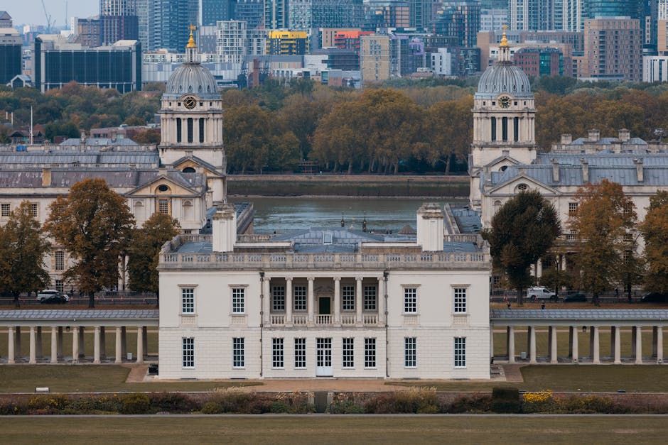 A wide-angle view of the Royal Naval College in Greenwich, London, showcasing its classical white façade with ornate architectural details, symmetrical towers with domed tops, and a series of colonnades along the front. The building is situated on a well-maintained grassy lawn with paved pathways leading towards it. The background features a city skyline with modern skyscrapers and high-rise office buildings, under a partly cloudy sky with patches of sunlight. The scene is set during daytime, with natural light illuminating both the historic structure and the urban backdrop. This image emphasizes the contrast between the historic Greenwich architecture and the contemporary cityscape, illustrating the area’s unique blend of heritage and modern development, which can be related to professional waste removal services handling clearance of historic sites or urban redevelopment projects in Greenwich.