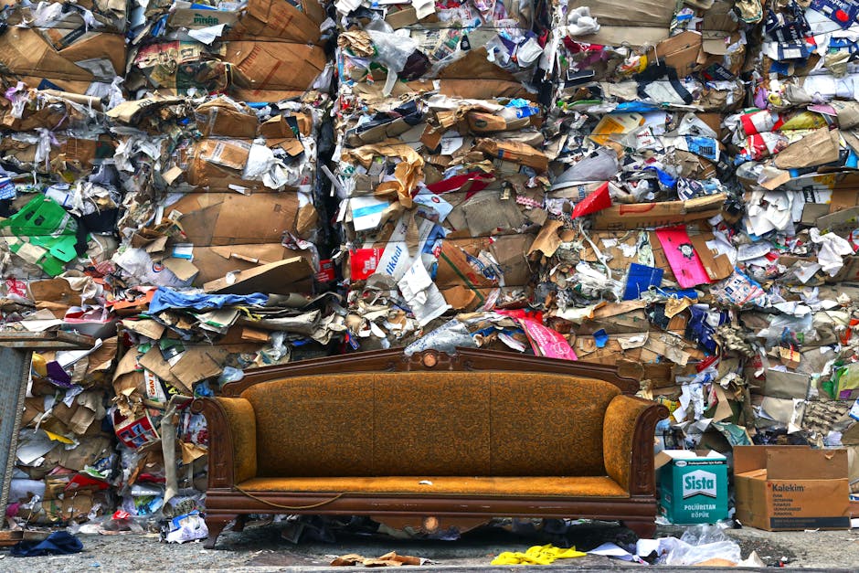 A vintage upholstered sofa with a wooden frame and carved detailing, featuring mustard-yellow fabric with an intricate floral pattern, is positioned in front of a large, densely packed wall of mixed waste materials. The waste comprises tightly compressed cardboard boxes, crumpled paper, and various packaging materials in shades of brown, white, blue, and red, creating a textured and chaotic background. The sofa is placed on an outdoor pavement area, with a few scattered items such as a cardboard box labeled 'Sista' and a yellow object nearby, indicating it may be part of a waste collection or clearance scene. The lighting appears natural, with daylight illuminating the cluttered environment, emphasizing the contrasting textures of the vintage furniture against the rough, dense layers of refuse. This scene subtly illustrates private rubbish removal or independent waste clearance efforts by Rubbish Removal Greenwich, highlighting the challenge of clearing large amounts of waste stored or accumulated in an urban or residential setting.