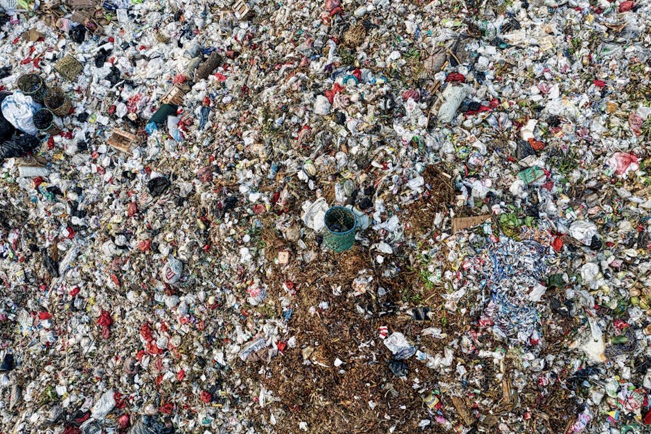 A large collection of discarded waste is piled on a paved area in front of a row of waste containers and a parked silver car with a visible license plate. The rubbish includes cardboard boxes, plastic bags, paper, and packaging materials, some of which are crumpled or torn. The waste is mainly contained in six different bins of varying sizes and colors, including grey with blue labels for mixed paper and cardboard, black for general waste, and a red bin likely for recyclable items. Some trash has spilled onto the surrounding pavement, forming a heap of mixed materials. In the background, there is a blue metal fence and a building with storefronts, one of which signage indicates a fish bar. A leafless tree is situated to the left of the waste without obscuring the scene, and the environment appears to be an urban street or parking lot, possibly associated with private waste collection services like Rubbish Removal Greenwich, who handle such waste clearance tasks efficiently.
