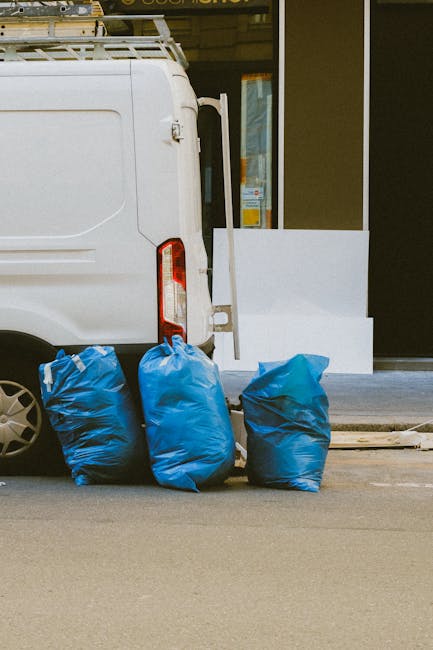 A white commercial van parked on a paved street, with its rear door slightly open, revealing part of the interior. Three large blue rubbish bags, made of durable plastic with wrinkled textures, are positioned on the ground next to the van, leaning against its rear wheel and side. The background shows a building with a glass window and a black door frame, suggesting an urban or commercial area. The scene appears to be part of a rubbish removal process, with the bags ready for collection, which is typical of private waste disposal services like those offered by Rubbish Removal Greenwich. The lighting is neutral, indicating daylight, and the environment appears clean and orderly, suitable for independent rubbish collection or on-site clearance activities involved in waste management, especially within Greenwich's local context.