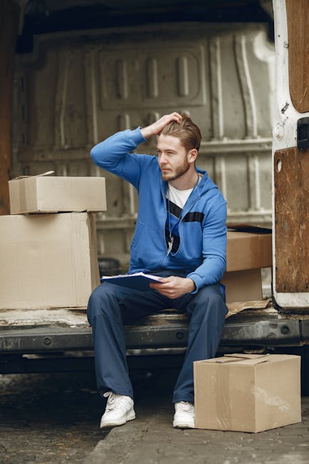 A young man with light brown hair and a beard, wearing a blue track jacket, white t-shirt, and blue trousers, is seated at the rear of an open delivery van. He is holding a clipboard in his lap while scratching his head with his right hand, suggesting a moment of thought or difficulty. The van's interior, visible behind him, shows a metallic, textured surface with vertical ridges. Surrounding him on the ground are several cardboard boxes, some stacked inside the van and others placed outside on the pavement. The boxes are plain, light brown, and vary in size, indicating a typical scenario of manual box-based rubbish or package collection. The scene appears to be set during daylight with natural light illuminating the area, contributing to a neutral and professional tone suitable for an article on rubbish removal or private waste handling services. The presence of the boxes and the location implies on-site clearance or logistics related to waste management, aligning with independent collection methods offered by services like Rubbish Removal Greenwich.