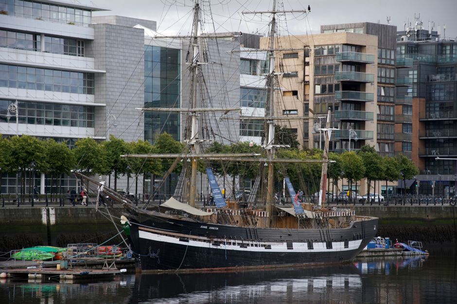 A historic tall ship with multiple masts and intricate rigging is docked along a modern urban riverfront in Greenwich. The vessel has a dark wooden hull with a white stripe running along its side and a deck lined with wooden planks, displaying a weathered yet well-maintained appearance. The masts are fitted with a complex network of ropes and sails, some furled, with the rigging spanning vertically and horizontally. The ship is moored next to a small floating platform with colorful kayaks and life buoys, secured with ropes. Behind the ship, a row of neatly trimmed green trees lines the promenade, separating the vessel from a backdrop of contemporary office and residential buildings characterized by glass facades, balconies, and a variety of neutral and muted tones. The water in the river appears calm, reflecting the ship and surrounding structures under an overcast sky, contributing to a subdued atmosphere typical of an industrial or urban waterfront. This scene exemplifies a contrast between historical maritime craft and modern city architecture, and while the image depicts a specific vessel, it broadly aligns with private or independent vessel moorings near urban settings, similar to alternative waste and rubbish removal contexts managed by companies such as Rubbish Removal Greenwich.