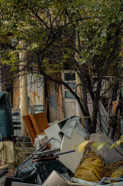 An outdoor scene featuring a pile of discarded household and building waste accumulated on a patch of ground beneath a tree with dense green foliage. The debris includes a variety of materials such as white appliances, wooden furniture pieces with orange-brown finishes, and broken construction materials, all partially covered by a grey fabric or tarp. The items are haphazardly stacked and appear to be in the process of disposal, indicative of an on-site clearance or private waste removal. Behind the waste, there is a backdrop of indistinct structures with worn, weathered surfaces, and some fencing or wire visible on the right side. Natural daylight illuminates the scene, highlighting the textures and colors of the debris and surrounding environment. This image demonstrates the type of rubbish removal handled by services like Rubbish Removal Greenwich, emphasizing the collection of bulky household items and construction waste typical of private clearance projects before disposal or recycling.