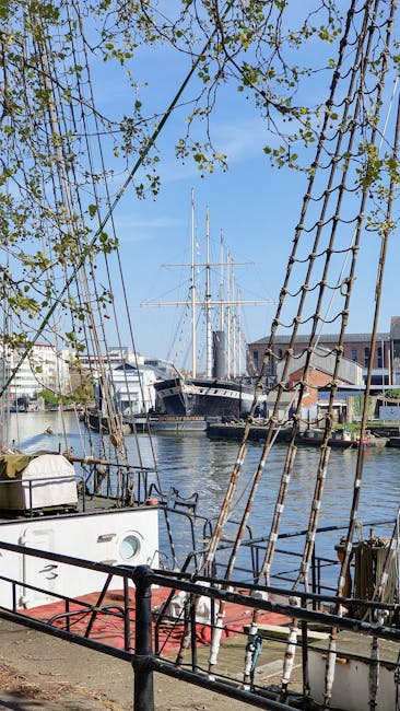 A historic tall ship with multiple masts and intricate rigging is docked along a modern urban riverfront in Greenwich. The vessel has a dark wooden hull with a white stripe running along its side and a deck lined with wooden planks, displaying a weathered yet well-maintained appearance. The masts are fitted with a complex network of ropes and sails, some furled, with the rigging spanning vertically and horizontally. The ship is moored next to a small floating platform with colorful kayaks and life buoys, secured with ropes. Behind the ship, a row of neatly trimmed green trees lines the promenade, separating the vessel from a backdrop of contemporary office and residential buildings characterized by glass facades, balconies, and a variety of neutral and muted tones. The water in the river appears calm, reflecting the ship and surrounding structures under an overcast sky, contributing to a subdued atmosphere typical of an industrial or urban waterfront. This scene exemplifies a contrast between historical maritime craft and modern city architecture, and while the image depicts a specific vessel, it broadly aligns with private or independent vessel moorings near urban settings, similar to alternative waste and rubbish removal contexts managed by companies such as Rubbish Removal Greenwich.