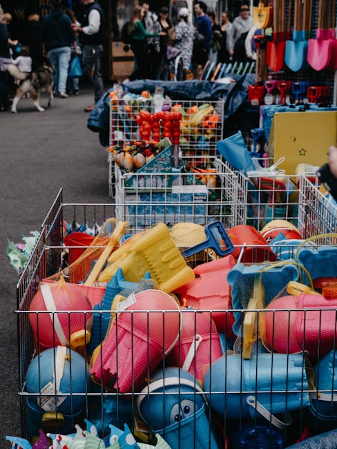 The image shows multiple metal wire baskets filled with colorful plastic beach toys, including buckets, spades, and water squirters, stacked and arranged on the pavement of an outdoor market or fairground. Some toys are yellow, pink, blue, and red, with smooth, shiny textures and rounded edges. Behind the baskets, there are other stalls and displays, with a variety of items possibly for sale, and a crowd of people browsing in the background. The scene appears to be well-lit with natural daylight, creating a bright and lively atmosphere. This setting suggests an environment where various goods, including children’s toys, are being sold or exchanged, potentially related to an outdoor or community event. The presence of toys and market activity may relate to private or third-party waste handling services like those offered by Rubbish Removal Greenwich, specifically supporting the clearing of small waste items from such venues or events in the Greenwich area.