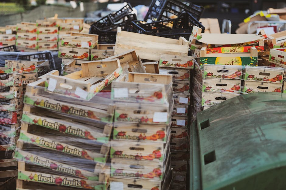 A large outdoor collection of empty wooden fruit crates branded with 'fresh fruits' stacked in multiple rows, with some crates slightly tilted or leaning against each other. The crates are light-colored wood with a smooth finish, featuring cut-out handles on the sides and printed labels showing images of various fresh fruits, primarily red apples and oranges, along with the text 'fresh fruits' in a green and red font. In the background, there are several black plastic crates piled haphazardly, and a green wheelie bin with a textured surface and rectangular ventilation slots near the top right corner. The scene appears to be set on a paved surface, possibly a warehouse or alleyway, under natural daylight, with a focus on the clutter of discarded or stored packaging materials. This image relates to the context of rubbish removal services, showcasing types of waste that might be collected during private on-site clearance or alternative waste handling processes, with the presence of various materials indicating an effort to manage waste in an organized and responsible manner.