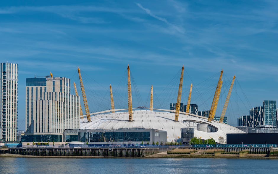 A wide-angle view of the distinctive London Olympic Stadium, featuring its white, textured dome roof supported by yellow structural masts and cables. The stadium is situated along a waterfront area, with the surrounding environment including modern high-rise office buildings with glass and steel facades on the left side of the image. The sky above is partly cloudy with patches of blue, casting natural light that highlights the stadium's architectural details. The foreground shows a paved promenade with a railing, and a water body extends across the bottom edge of the image, emphasizing the stadium's prominent position in the urban landscape. This scene represents an example of infrastructure that might be associated with large-scale construction or renovation projects, which occasionally generate construction waste requiring specialist rubbish removal services such as those offered by Rubbish Removal Greenwich, especially when managing territory clearance or site preparation for city development projects.