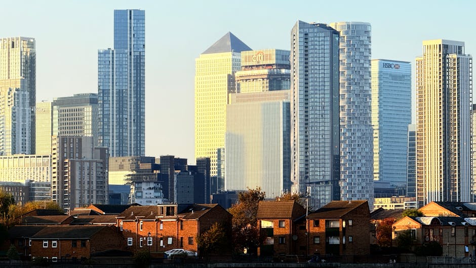 A cityscape featuring a mix of tall modern skyscrapers and smaller brick residential buildings situated in the foreground. The skyscrapers are composed of glass and steel, exhibiting reflective surfaces and contemporary architectural styles with various heights, shapes, and facades, some with visible company logos such as HSBC and Citi near the tops. The brick houses in the foreground have pitched roofs, with some chimneys and small windows visible, and are arranged in a row along a street or residential area. The overall lighting indicates late afternoon sunlight, casting a warm glow on the buildings and creating subtle shadows. The scene is set against a clear sky, emphasizing the contrast between the dense commercial high-rises and the lower residential structures. This urban environment illustrates a typical city boundary, where private residential areas meet the towering commercial skyline, relevant to discussions about urban development, property management, and the varied waste disposal needs of such mixed-use environments, as coordinated by waste management services like Rubbish Removal Greenwich.