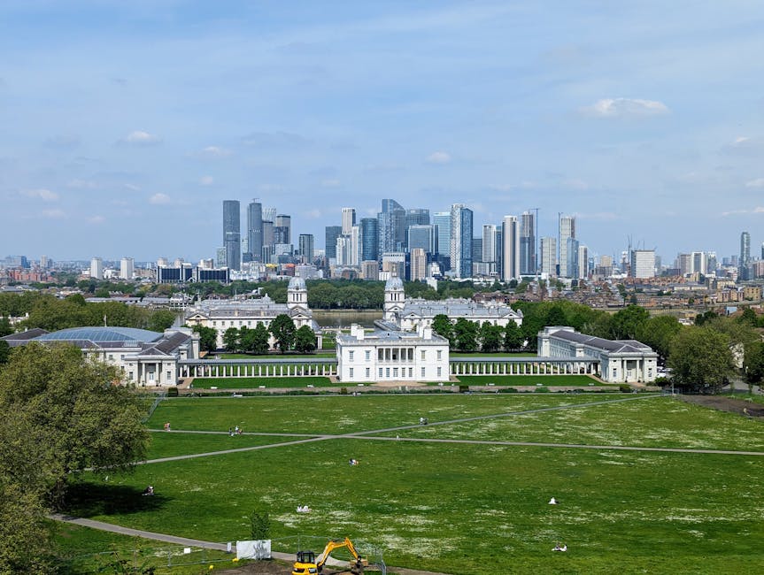 A wide-angle view of a large historical white building with classical architectural features, including columns and domed roofs, situated in a lush green park with well-maintained lawns and paved walking paths. The park is populated with a few small groups of people relaxing and walking. In the foreground, there's a small yellow excavator positioned near a construction area. Behind the building, there are dense trees and a modern city skyline with numerous high-rise office and residential towers made of glass and steel, reflecting sunlight against a partly cloudy sky. The scene showcases the contrast between the open, green public space and the surrounding urban environment, illustrating a typical context where private waste handling and rubbish removal services, such as those offered by Rubbish Removal Greenwich, could support the maintenance and clearance of waste generated in such areas.