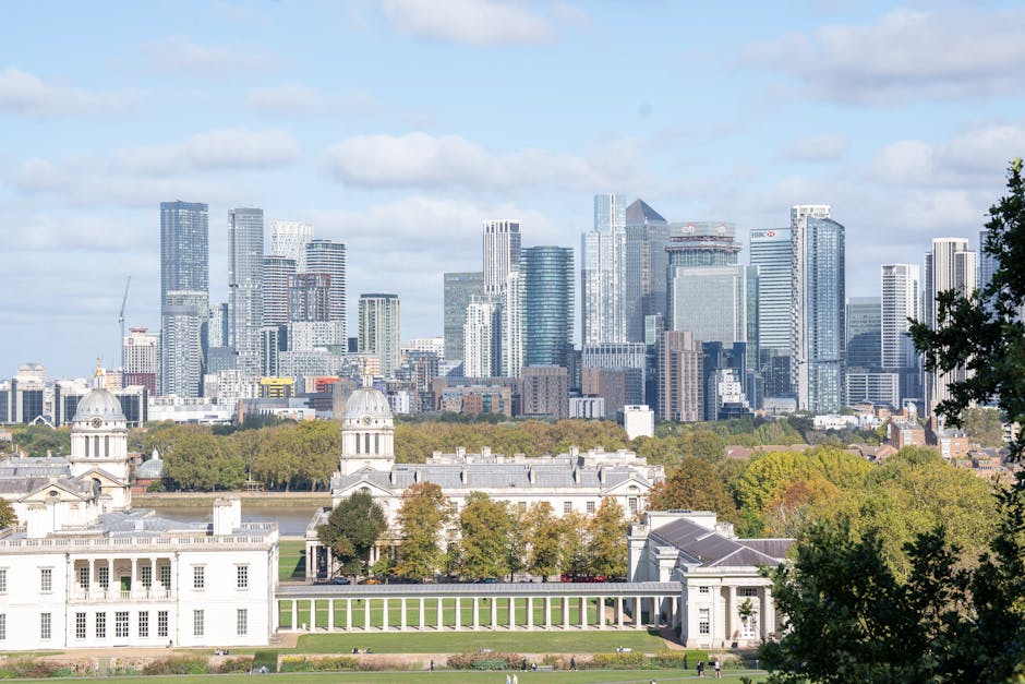A panoramic view of a modern city skyline featuring numerous high-rise office buildings and skyscrapers with reflective glass facades under a clear blue sky. In the foreground, there is a lush area of greenery with dense trees and well-maintained grass, providing a natural contrast to the urban landscape. The trees vary in species, some with broad leaves and others with needle-like foliage, creating a vibrant and textured scene. The city buildings, some under construction with cranes visible, occupy the middle and background sections of the image, while the foreground offers an open, park-like environment. The overall atmosphere is bright, suggesting daytime, with natural sunlight illuminating both the trees and the city structures. This scene illustrates the relationship between urban development and green spaces, relevant for discussions on private waste management and alternative rubbish removal methods in city environments, as presented on Rubbish Removal Greenwich's page about Greenwich residents' clearance steps.