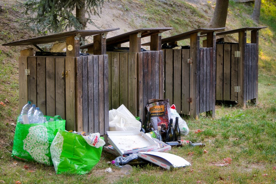 A narrow outdoor pathway bordered on the left by a weathered wooden fence and on the right by dense green foliage and bushes. The ground is composed of dirt and patches of grass, with some areas appearing muddy. Several discarded wooden pallets are stacked horizontally against the hedge on the right, with some leaning at slight angles, and a white plastic bag or sheet draped over part of the pallets. In the background, tall trees with bright green leaves cast dappled sunlight over the scene, creating a play of light and shadow. Beyond the trees, the rooftops of residential buildings and a few taller high-rise structures are visible against a clear blue sky, suggesting the setting is in a suburban or urban fringe area. The scene indicates an informal or ongoing outdoor clearance, potentially managed by private waste removal services such as Rubbish Removal Greenwich, to clear discarded furniture or garden waste from the site in preparation for proper waste disposal or recycling, aligning with alternative waste handling practices.